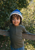 Child wearing Denmark Beanie Blue & Cream  outdoors with greenery in the background – kids striped knit beanie with pointed crown – Acorn Kids 