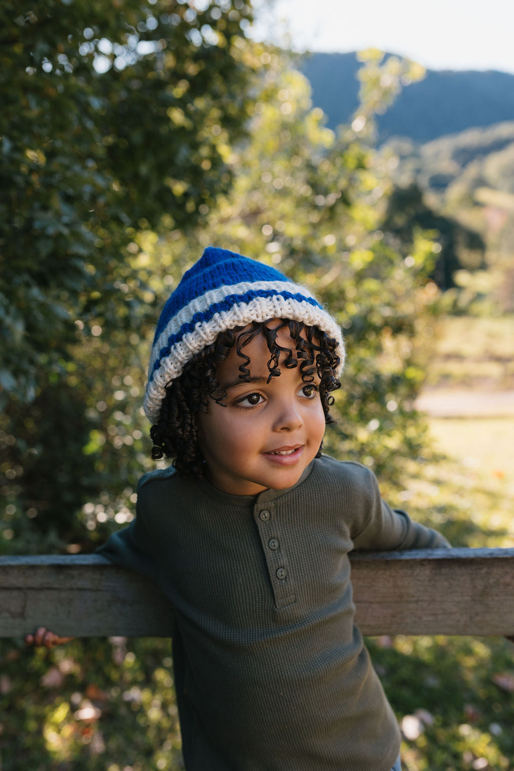 Child wearing Denmark Beanie Blue & Cream front view standing outdoors with greenery in the background – kids striped knit beanie with pointed crown – Acorn Kids 